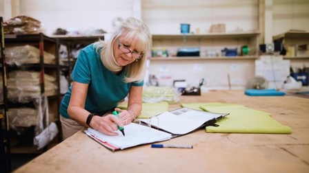 Person mit blonden Haaren und Brille mittleren Alters steht in einer Schneiderei und markiert Blätter einer geöffneten Arbeitsmappe während ein grüner Stoff neben ihr auf der Arbeitsfläche liegt. 