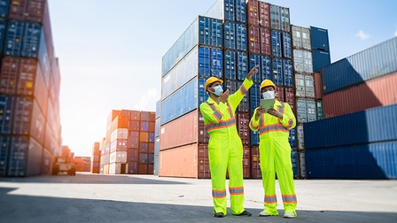 Zwei Personen in gelber Arbeits- und Schutzbekleidung mit gelben Schutzhelm sowie Mund-Nasen-Maske, rundherum stehen viele Container in den Farben blau, rot und weiß.