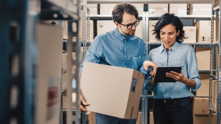 Zwei Personen in Gang zwischen Regalen voller Kartonagenboxen stehend und auf Tablet blickend, das eine der Person in Händen hält, anderer Person hält großes Paket unterm Arm