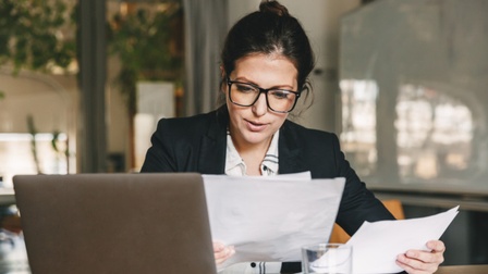  Person mit Brillen sitzt an Tisch und sichtet Dokumente, aufgeklapptes Notebook und Wasserglas auf Tisch platziert