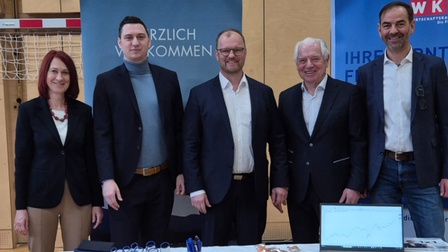 Fünf Personen stehen gemeinsam hinter einem Infostand bei einer Berufsmesse in einem Turnsaal. Im Hintergrund sind Roll-ups und ein Willkommensbanner zu sehen, auf dem Tisch liegen Informationsmaterialien und ein Laptop.