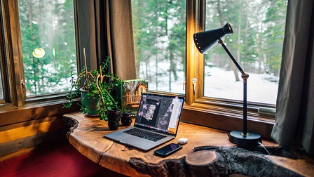 Ein Laptop steht auf einem Holz-Schreibtisch bei einem Fenster und eine Schreibtischlampe leuchtet darauf