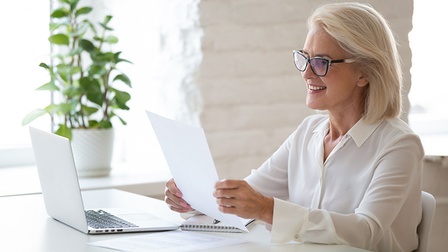 An Schreibtisch sitzende lächelnde Person mit Brillen blickt auf Blatt in Händen, am Tisch aufgeklappter Laptop, im Hintergrund Zimmerpflanze