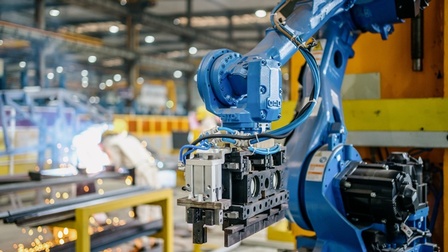 Blick auf einen blauen Roboterarm in einer Fabrik. Im Hintergrund ist eine Industriehalle mit gelben und metallenen Elementen zu sehen.