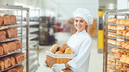 Lächelnde Person in weißem Kochgewand mit Haube hält Korb mit Brotlaiben in Händen