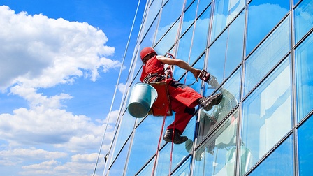 Person in roter Arbeitsausrüstung mit Helm hängt an Seilen und reinigt Fensterfassade