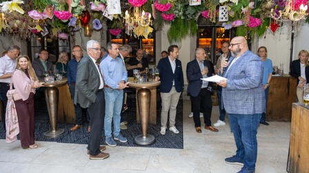 Mann spricht mit Mikrofon zu einer Gruppe bei einer Veranstaltung mit festlicher Dekoration und Stehtischen.