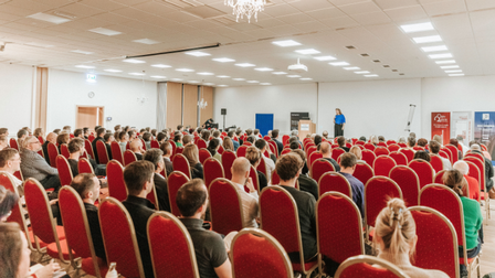 Großer Veranstaltungssaal mit zahlreichen Besucher:innen auf roten Stühlen, die einer Vortragenden auf der Bühne zuhören; im Hintergrund stehen Roll-ups und eine Präsentationsfläche.