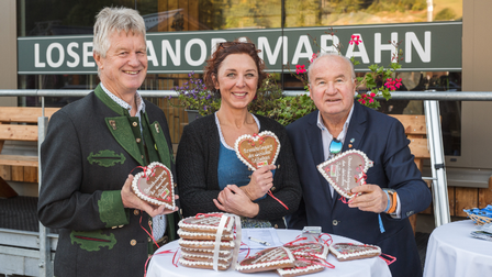 Zwei Männer und eine Frau in Tracht stehen vor einem Stehtisch mit Lebkuchenherzen, jeder hält ein Herz in der Hand
