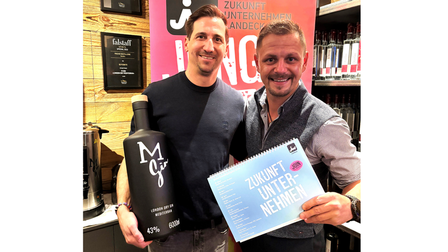 Zwei Personen stehen nebeneinander in einem Geschäft oder Showroom mit Holzregalen und Flaschen im Hintergrund. Beide lächeln in die Kamera. Der Mann links hält eine große schwarze Flasche mit der Aufschrift „M Gin“ in den Händen. Der Mann rechts hält einen blauen Block oder ein Heft mit der Aufschrift „Zukunft Unternehmen“. Im Hintergrund sind Regale mit Flaschen sowie Schilder und Auszeichnungen an der Wand zu sehen. Die Szene wirkt wie eine Präsentation oder ein gemeinsamer Termin im geschäftlichen Umfeld.