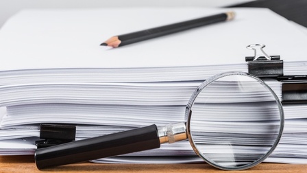 Detailansicht eines Aktenstapels mit Klammern, darauf liegend ein Bleistift, Vergrößerungslupe angelehnt