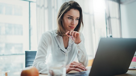 Person stützt Hand mit Brille an Kinn und blickt auf aufgeklapptes Notebook