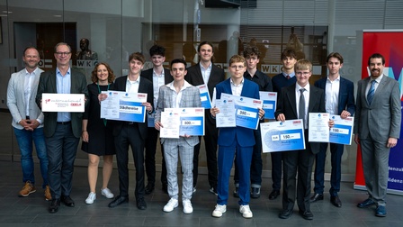 Eine Gruppe von mehreren Personen steht in zwei Reihen in einem Innenraum vor einer Glaswand mit WKO-Beschriftung. Mehrere Personen halten Urkunden und großformatige Schecks mit Beträgen wie „150“, „350“ und „500“. Die Personen tragen formelle Kleidung wie Anzüge und Hemden. Links und rechts stehen jeweils weitere Personen ohne Urkunden