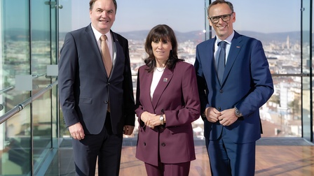 Porträt von drei Personen nebeneinander vor einer Fensterfront mit Blick über eine Stadt
