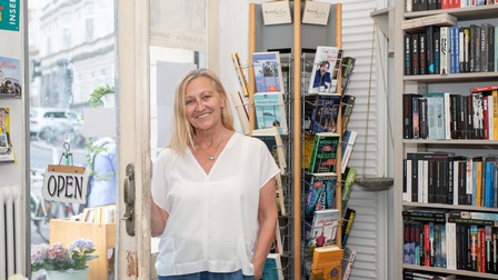 Porträt einer lächelnden Person mit offenen, hellen Haaren, die in einem Geschäft in der geöffneten Tür steht. Hinter der Person sind Regale mit Büchern