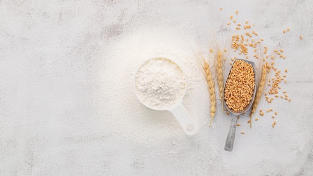 Küchenmesslöffel voller Mehl, daneben Metallschäufelchen voller Weizenkörner und goldgelbe Weizenähren in Vogelperspektive