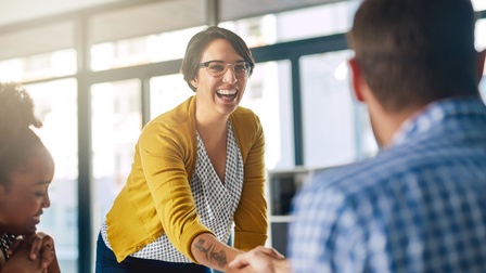 Lächelnde Person mit Brille in gelber Jacke mit Tätowierung am Unterarm schüttelt anderer Person die Hand