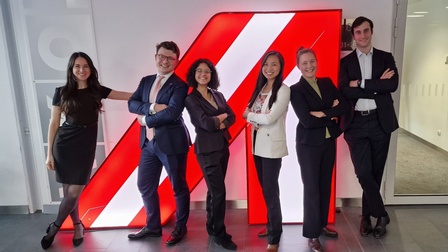 Gruppenfoto von sechs Personen in Bürokleidung, die nebeneinander vor einem großen rot-weiß-roten A in einem Gang stehen