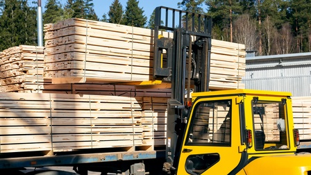 Auf einer erhöhten, blauen Metallplatte stehen viele Stapel heller Holzbretter. Vor der Platte steht ein gelber Gabelstapler, der einen Stapel Holzbretter hochhebt. Im Hintergrund sind hohe, grüne Bäume