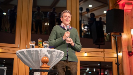 Eine Person in Trachtenkleidung steht vor einer Fensterfront und hält ein Mikrofon. Neben der Person steht ein Tisch mit Getränken. In der Fensterfront spiegeln sich ein paar Personen