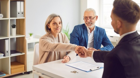 Zwei Personen sitzen an einem Tisch. Die linke Person schüttelt einer ihr gegenüber stehenden Person die Hand. Auf dem Tisch liegen ein Zettel auf einem Klemmbrett und ein Schlüssel