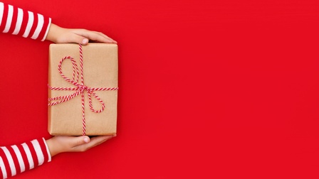 Auf rotem Hintergrund halten im linken Bildrand zwei Hände einer Person in rot-weiß gestreiftem Oberteil ein rechteckiges Geschenk, eingepackt in hellbraunem Geschenkspapier mit rot-weißem Geschenksband