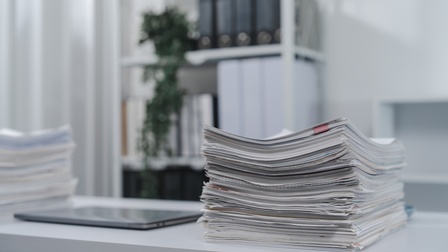 Auf einem weißen Tisch ist ein Dokumentenstapel. Daneben sind ein Tablet und ein weiterer Dokumentenstapel. Im Hintergrund ist ein Regal mit Ordnern