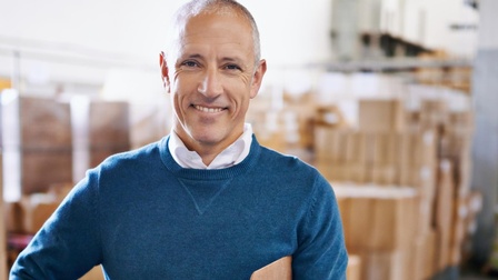 Person mit Pullover steht mit einem Klemmbrett und Kugelschreiber freudig in einer Lagerhalle