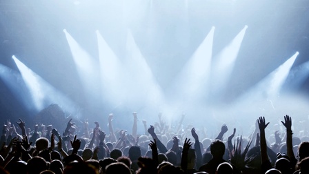 Viele Personen, Fans, stehen vor einer Bühne, die mit Scheinwerfern beleuchtet ist