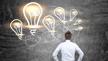 Eine Person ist von hinten fotografiert, sie steht vor einer grauen Wand mit weißen Sprenkeln. Auf der Wand sind Illustrationen Glühbirnen, bei denen die Lichtquelle grell hervorsticht. Sie sind mit strichlierten und gebogenen Linien verbunden. 