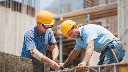 Zwei Personen mit gelben Schutzhelmen arbeiten an der Montage auf einer Baustelle, eine Person trägt Schutzverband um den Arm