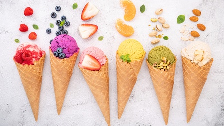 Auf hellem Untergrund liegen sechs Eistüten nebeneinander. In jeder Tüte ist eine Eiskugel mit einer anderen Farbe. Vor jeder Kugel liegt ein Obst oder eine Nuss, von links nach rechts Himbeeren, Blaubeeren, Erdbeeren, Mandarinen, Pistazien, Mandeln