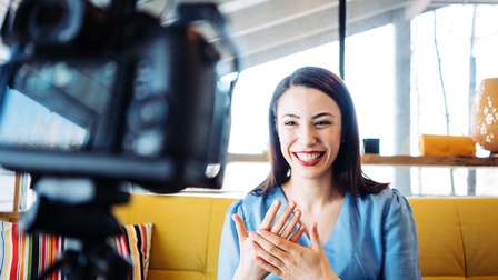 Lächelnde Person mit dunklen Haaren und roten Lippen strahlt in die Kamera, die im Vordergrund unscharf in Bild kragt