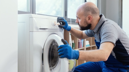 Eine Person mit Bart, Glatze, blauer Latzhose und Handschuhen kniet neben einer geöffneten Waschmaschine und dreht an einem Knopf an der Waschmaschine. Im Hintergrund steht ein Regal mit Waschmitteln
