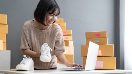Lächelnde Person steht an Tisch, blickt auf aufgeklapptes Notebook und hält in einer Hand weißen Sneaker, im Hintergrund gestapelte Kartonagen