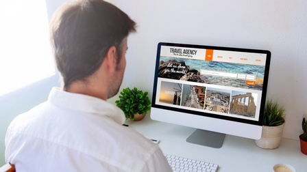 Rückenansicht einer Person, die an einem Schreibtisch sitzt. Vor der Person sind eine Tastatur und ein Computerbildschirm. Auf dem Bildschirm sind verschiedene Reisebilder. Darüber steht Reiseagentur