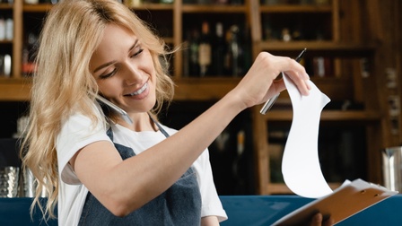 Person mit langen blonden Haaren und Schürze telefoniert mit einem Smartphone in der Schulter eingeklemmt und steht vor einer Schank und blickt auf Dokumente auf einem Klemmbrett