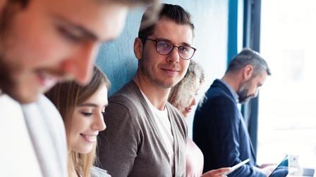 Mehrere Personen nebeneinander stehend, in verschiedene Smartdevices blickend, eine Person mit Brillen blickt in Kamerarichtung