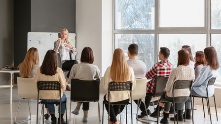Mehrere Personen in Rückenansicht sitzen auf Stühlen in Raum mit großen Fensterfronten und blicken auf Person vor Whiteboard, die Bürste in Hand hält und darauf deutet