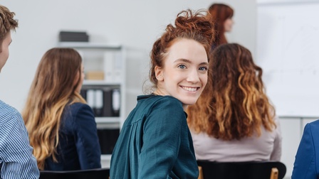 Lächelnde Person in Unterrichtssituation dreht sich nach hinter, Rückenansichten anderer Personen ringsum