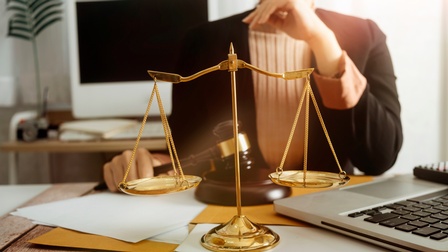 Eine Person sitzt an einem Tisch. Mit der rechten Hand hält sie einen Richterhammer. Vor ihr liegen Unterlagen. Auf dem Tisch steht ein aufgeklappter Laptop und eine goldene Waage