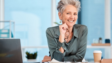 Person mit kurzen grauen Haaren, grüner Bluse und Ohrringen hält eine Brille in der Hand, während sie freudig an einem Schreibtisch abgestützt ist, daneben steht ein Laptop sowie ein Kaffeebecher sowie befinden sich Unterlagen am Tisch