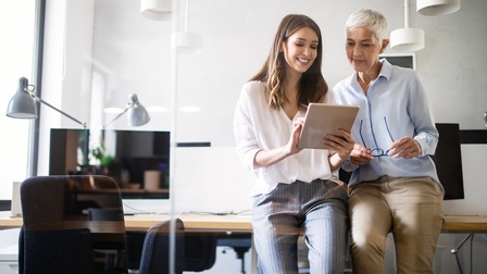 Personen in Businesskleidung stehen angelehnt bei einem Schreibtisch und blicken freudig auf ein Tablet, eine der beiden hält dabei eine Brille in den Händen