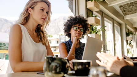 An einem Tisch sitzen sich vier Personen gegenüber. Von den Personen rechts sind nur die Hände im Bild. Die anderen Personen blicken in den rechten Bildbereich. Vor der linken Person steht ein aufgeklappter Laptop.