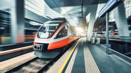 Auf Gleisen fährt ein einstöckiger, rot-grauer Zug. Rechts neben den Gleisen ist ein Bahnsteig