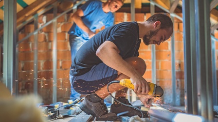 Seitliche Aufnahme einer auf einem Boden hockenden Person, die in der rechten Hand ein technisches Gerät mit Kabel hält, von dem Funken abgehen. Die Person blickt auf das Gerät. Im Hintergrund steht eine weitere Person