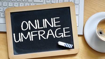 Auf einer Schiefertafel steht in Versalien das Wort Onlineumfrage. Darauf liegt ein Stück Kreide. Unter der Schiefertafel ist die Tastatur eines Laptops. Daneben in rechter Bildhälfte ist eine Untertasse und eine Tasse mit Kaffee
