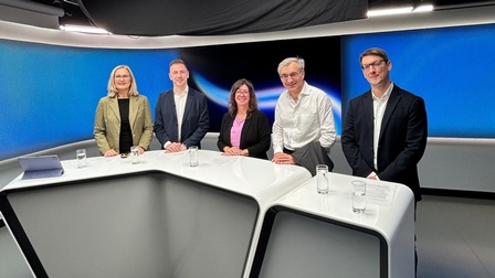 Fünf Personen stehen in einem modernen TV-Studio hinter einem Studiotisch.