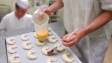 Teig ist zu Croissants geformt worden, diese sind auf einem Backblech mit Backpapier platziert und werden mit einer Flüssigkeit von einer Person mit Schutzbekleidung bestrichen, bereit für den Backvorgang
