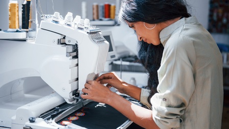 Person in Seitenansicht sitzt an Stickmaschine und bedient diese, unter Maschine schwarzer Stoff mit bunten gestickten Ornamenten